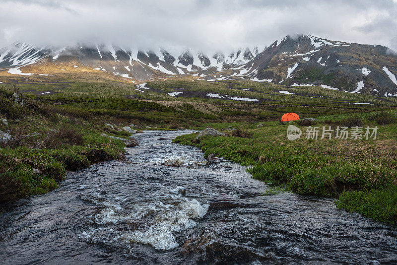 橙色帐篷靠近清澈的小溪，在低低的雨云中可以看到山脉。戏剧性的高山景观与山溪和雪山在灰色的低云。冻土带阴森的景色。