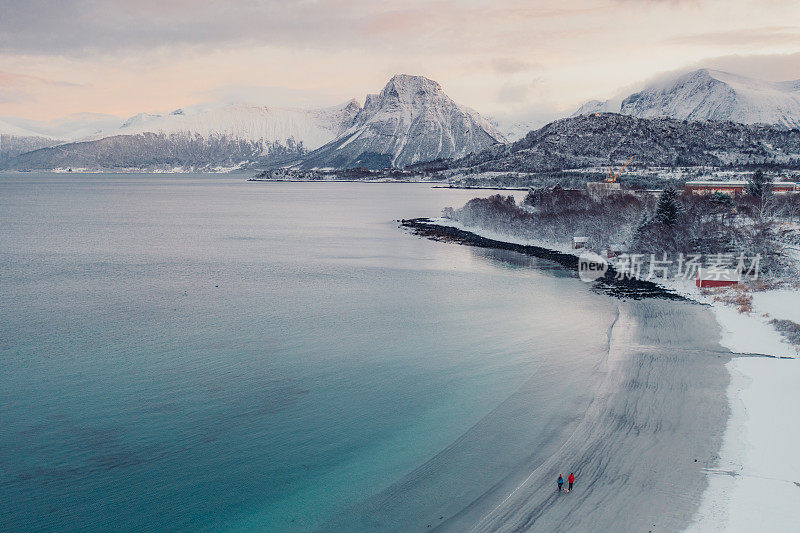 航拍图像的女人和男人走在风景秀丽的turqouse海滩在雪与山景在挪威