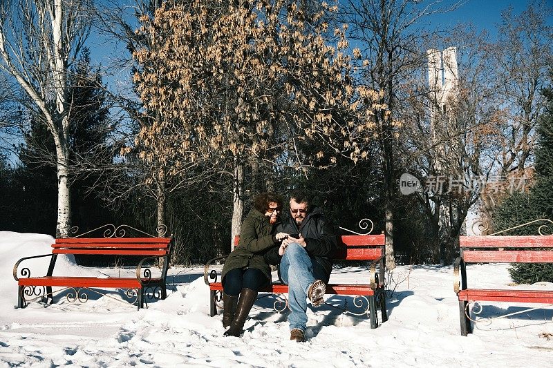 一对成熟的夫妇坐在冬天公园的长椅上玩手机