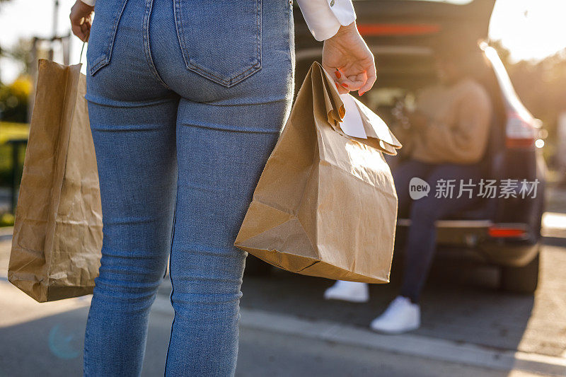 后视图的年轻女子背着一个纸袋外卖食品的订单，走向她的朋友正在等待的汽车