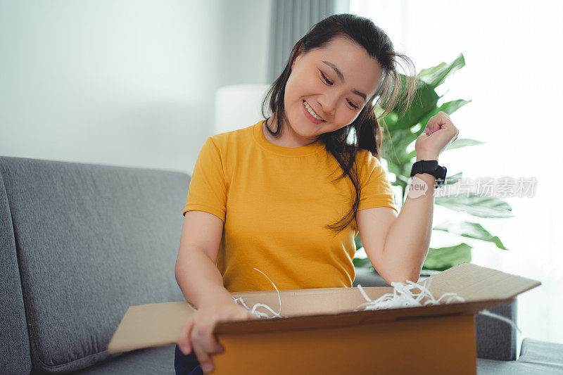 亚洲女人打开纸箱，开心兴奋地看着里面，坐在家里客厅的沙发上。