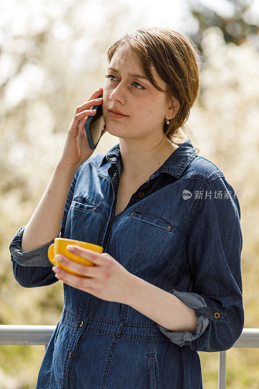 年轻女子站在阳台上，一边喝咖啡一边打电话