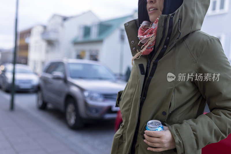 一个穿着夹克的女人站在人行道上，手里拿着一罐果汁。冰岛