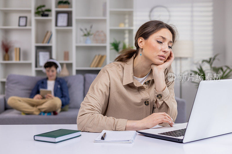 疲惫的女商人远程在家用笔记本电脑工作，用手抱着头，敲击键盘，编程，母亲在工作，孩子在后台