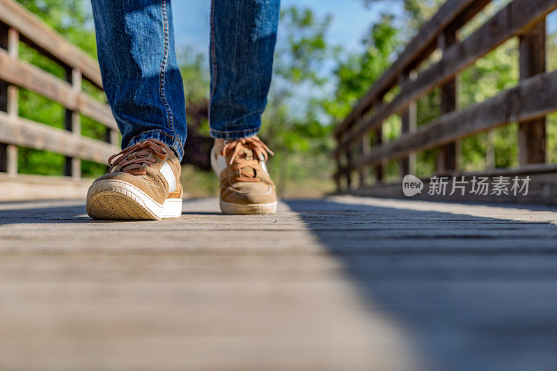 一个穿着牛仔裤和运动鞋的男人在一座自然公园的木桥上行走的部分照片。