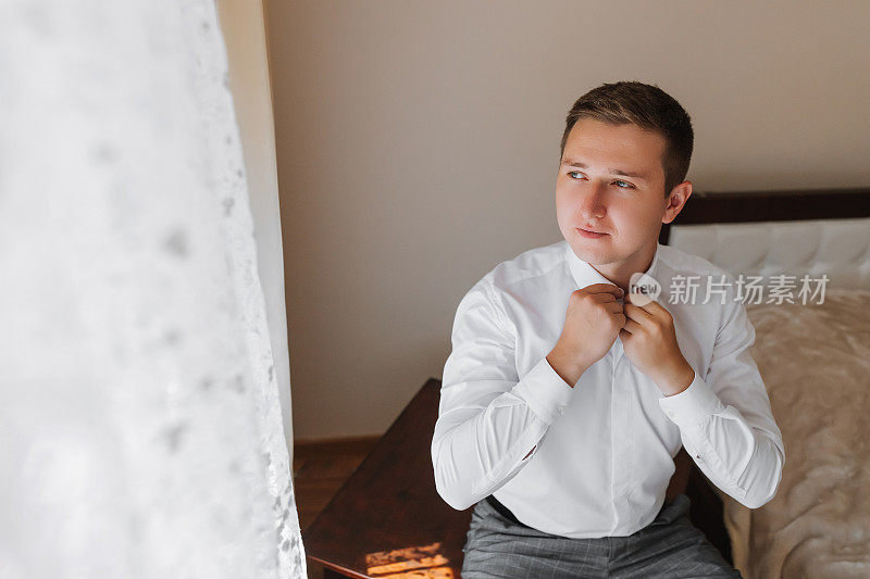 一位英俊的年轻男子在婚礼前摆出新郎的姿势，整理着衬衫的领子。新郎正在一幢大楼里为婚礼做准备