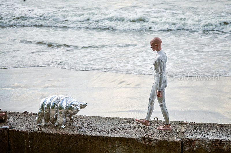 一个光头的女孩穿着未来派风格的衣服，带着玩具缓步动物在大海背景下散步