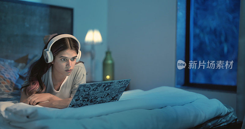 受惊吓的印度年轻成年z世代女性躺在床上，在家室内看恐怖电影。美丽的害怕的女孩用耳机在笔记本电脑的网络摄像头屏幕上看恐怖电影，享受深夜的时光