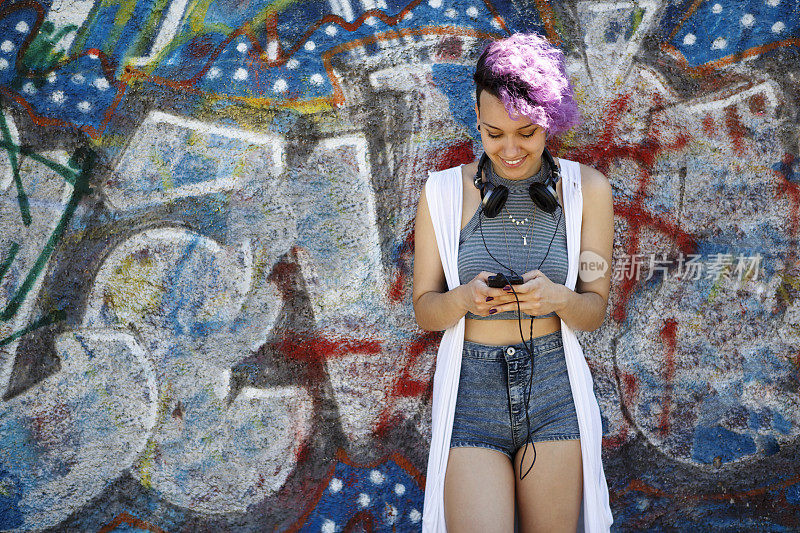 微笑的年轻时髦的女孩用智能手机对涂鸦墙