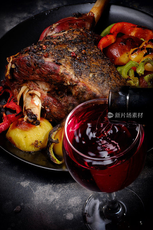 烤羊小腿配烤蔬菜，配红酒杯。垂直的图像。