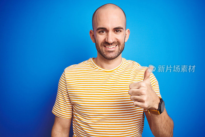 年轻的秃顶男子与胡子穿着休闲t恤在蓝色孤立的背景做快乐的拇指手势与手。赞许的表情看着镜头，表示成功。