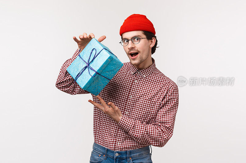 一个小伙子试着为女朋友准备了一份有趣的礼物，摇晃着装着礼物的盒子，想知道里面是什么，看着神秘而狡猾的相机，戴着红色的无檐小便帽，白色的背景