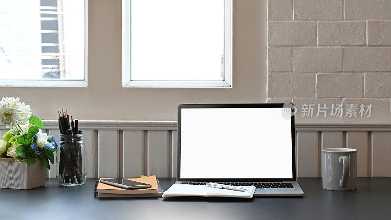 白色的黑屏笔记本电脑放在现代的办公桌上，包括盆栽、咖啡杯、笔记本、笔筒和智能手机。舒适的工作场所的概念。