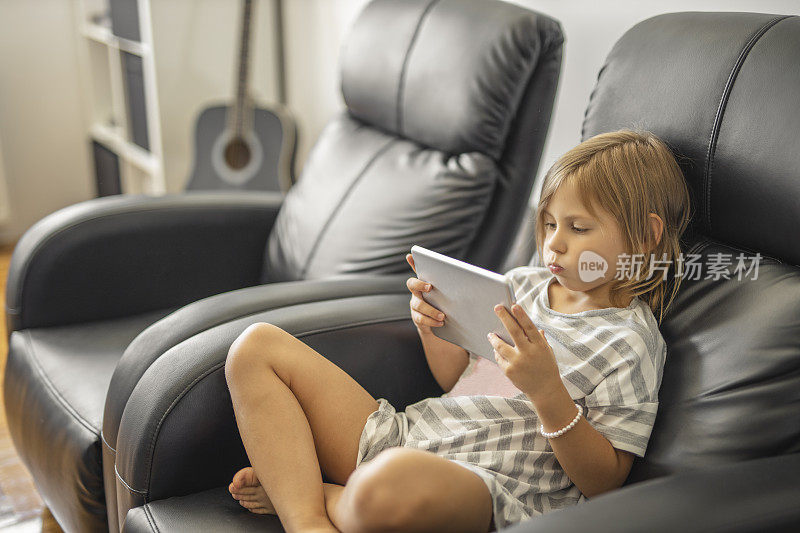 担心的小女孩穿着睡衣，坐在扶手椅上，在家里使用数码平板电脑