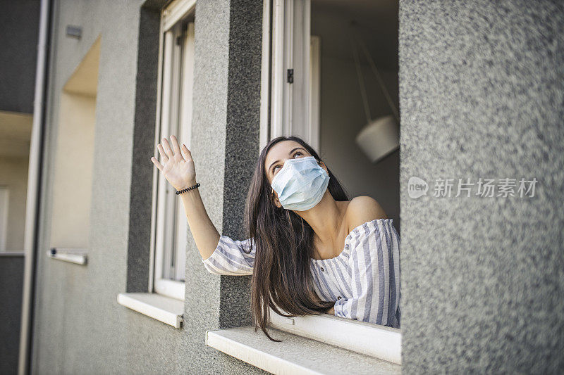 一名年轻的成年女子倚靠在一扇打开的窗户上，透过窗户向外看，并向邻居们挥手致意，而此时她正在家中休息，因为冠状病毒被隔离