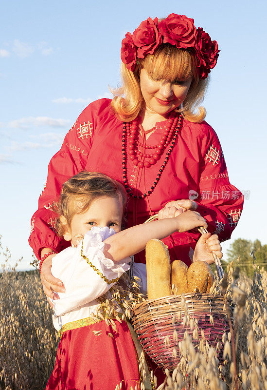 一个女孩拿着一个柳条篮子，里面放着新鲜的面包，背景是一片燕麦田。一个小孩，一个5岁的女孩，往篮子里看。穿着红裙子，头上戴着花环。日落。