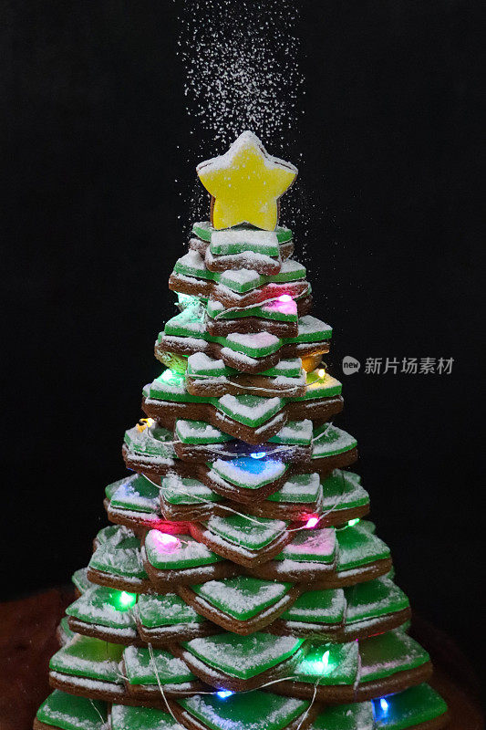图像糖霜糖筛在圣诞树上堆叠的姜饼星饼干与绿色方糖糖霜，装饰着发光的仙女灯，彩色糖果巧克力，黄色糖霜星饼干顶部，黑色背景