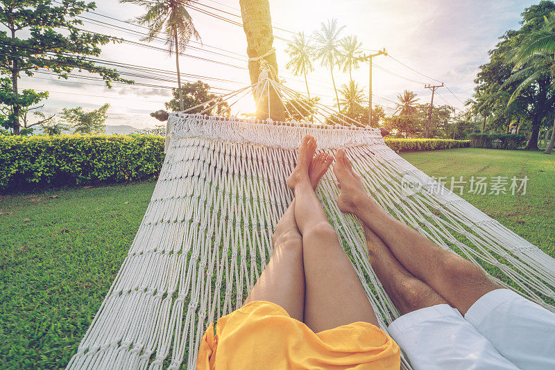 个人视角的夫妇放松在吊床上，脚的观点。一对夫妇的观点，从吊床在热带花园日落，赤脚。人们旅游热带气候概念。