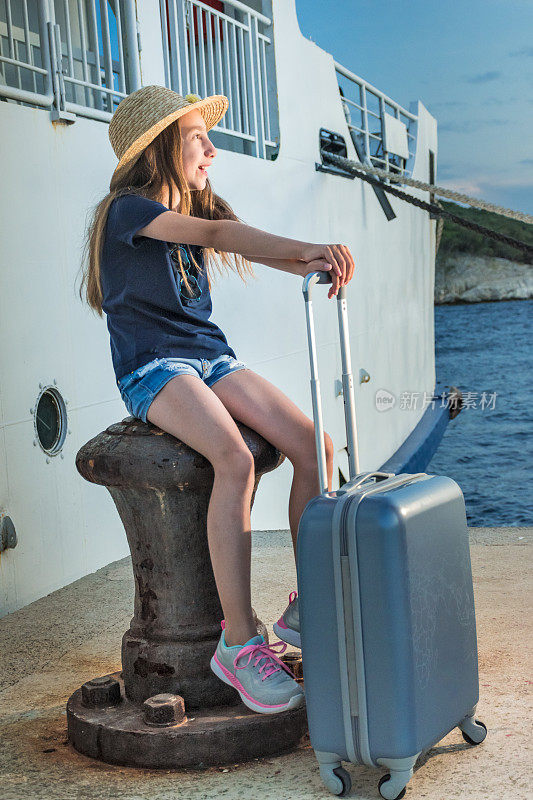 快乐的女孩拿着手提箱坐在码头上，在夕阳下等待渡船之旅