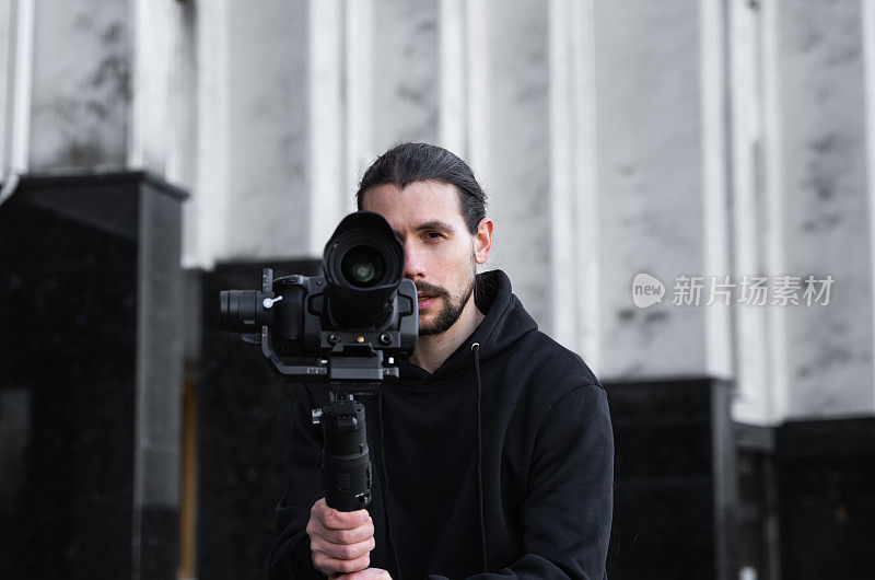 年轻的专业摄像师手持专业摄像机，安装在三轴框架稳定器上。专业设备有助于制作高质量的视频而不摇晃。一个穿着黑色连帽衫的摄影师正在录像。