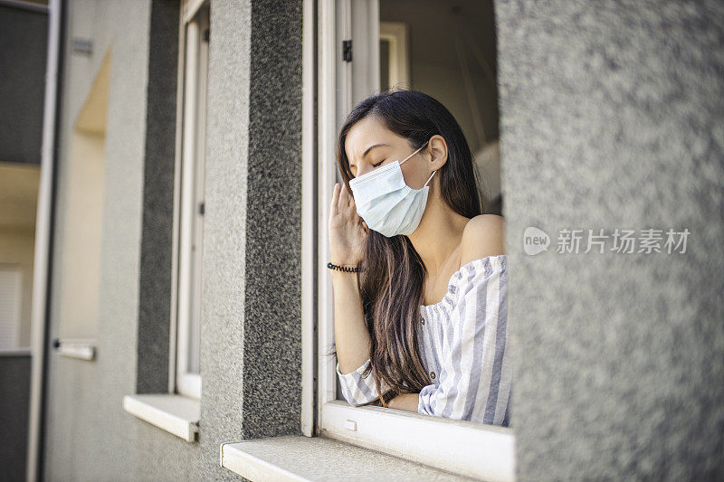 一名年轻的成年女子在隔离期间因冠状病毒感染而头痛并戴着医用口罩，她靠向打开的窗户向外看