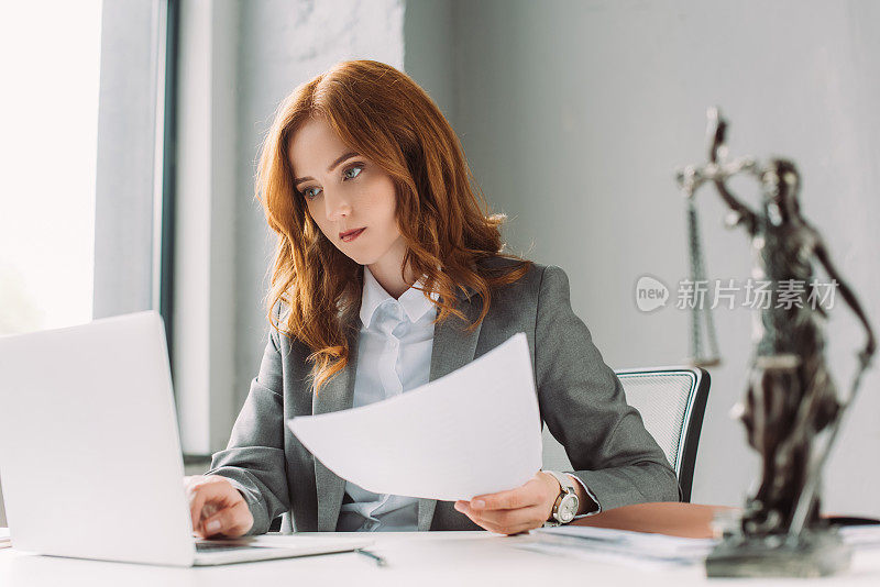 红发女律师与纸看笔记本电脑在工作场所，模糊的themis雕像在前景