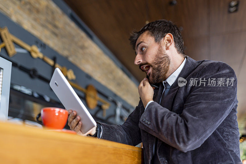 一个微笑的商人正在现代咖啡店里与商业伙伴通电话。他正坐在他的数字平板电脑，享受着一杯咖啡。