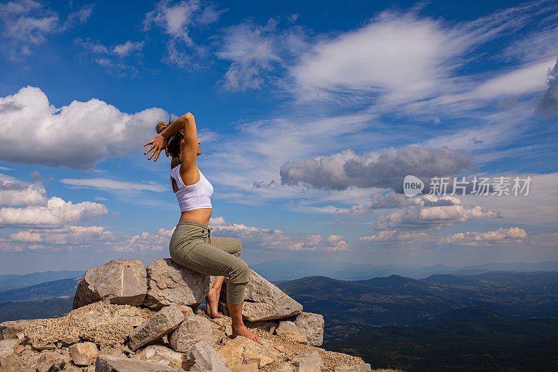 中年妇女，在岩石山顶保持平衡，同时做牛脸瑜伽姿势