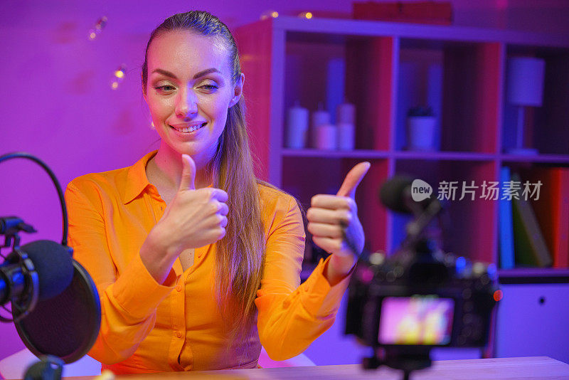 在一间紫色灯光下的摄影棚里，一位年轻可爱的女性博主，金发碧眼，身穿黄色衬衫，正对着摄像机，竖起大拇指