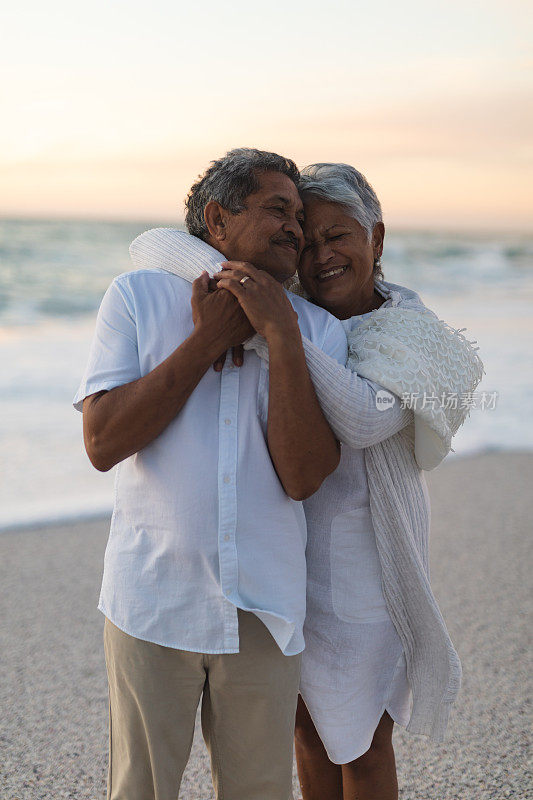日落时分，站在海滩上，一个快乐的混血年长女子从后面拥抱着男人