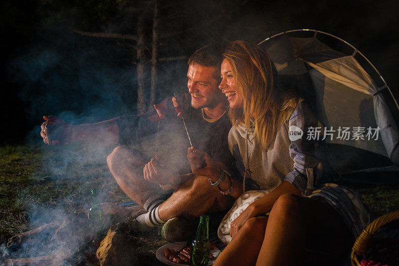 年轻的夫妇在篝火上烤香肠，在黄昏的森林山喝啤酒