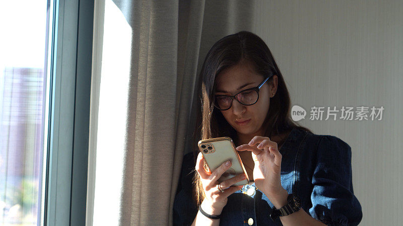 成功的年轻女子戴着眼镜，靠窗边用笔记本电脑聊天。使用IT进行写作和搜索。快乐微笑的布伊桑女士工作在智能手机浏览互联网。教育技术的概念
