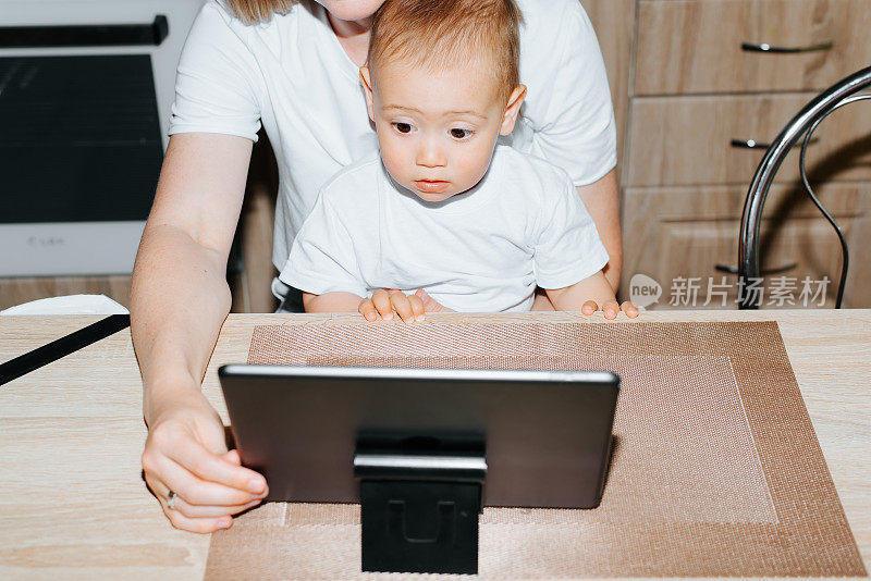 妈妈和孩子坐在家里的厨房里，一起使用平板电脑，看教育视频或动画片。小男孩情绪激动地看着小玩意儿。为人母，为人父母，照顾孩子。裁剪