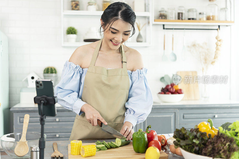 亚洲女性美食博主在智能手机摄像头前烹饪沙拉，同时在家里的厨房录制vlog视频和直播。