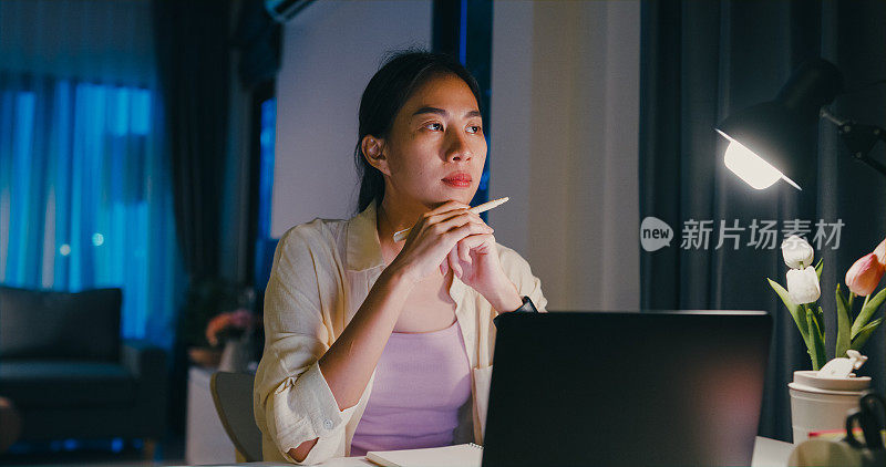 年轻的亚洲女性穿着正装衬衫坐在桌子前与笔记本电脑和文书工作想主意使紧急报告会议加班到晚上在家。