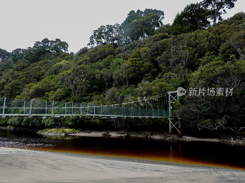 悬索式人行桥横跨一条充满单宁酸的入海河流。拉基拉赛道，斯图尔特岛，新西兰。