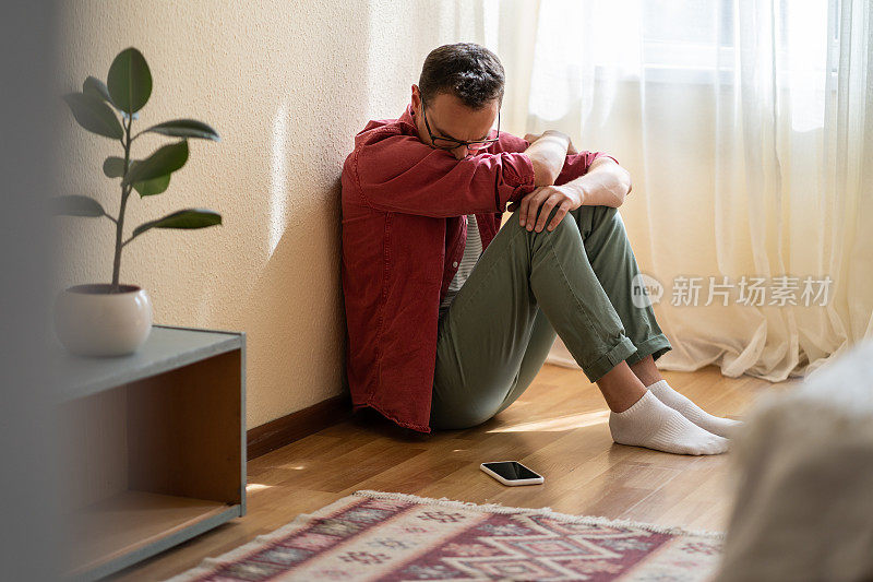 沮丧的白人男子坐在公寓的地板上，悲伤地看着手机等待电话