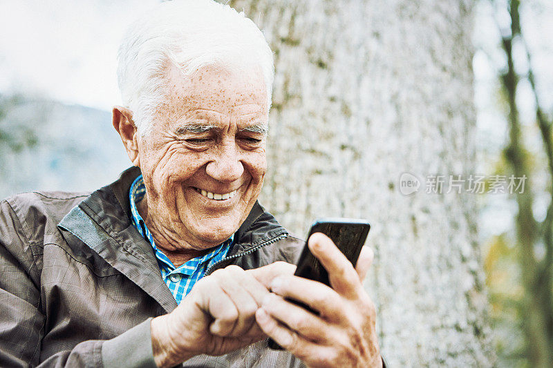 在户外的一棵树旁，一位高兴的老人微笑着用手机发短信