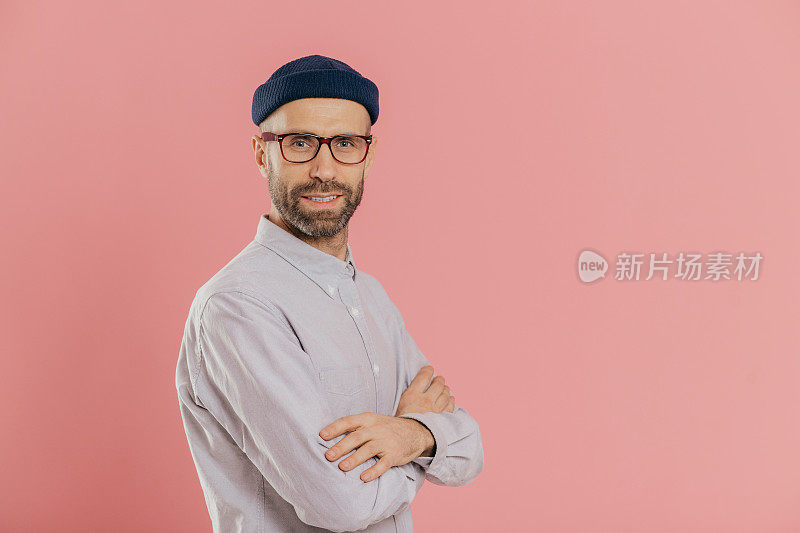 工作室拍摄自信的蓝眼睛大胡子男子，戴着时髦的帽子和白衬衫，双臂交叉，模特在粉红色背景上，左侧有广告内容的自由空间