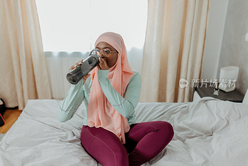 戴着头巾的年轻穆斯林妇女坐在床上，从运动瓶中取水补充水分