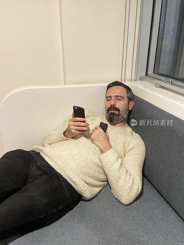 男子睡在沙发上，手里拿着智能手机和电视遥控器。睡觉的人