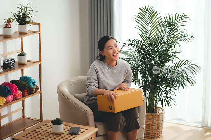 亚洲女人拿着纸盒，开心兴奋地坐在家里客厅的扶手椅上。