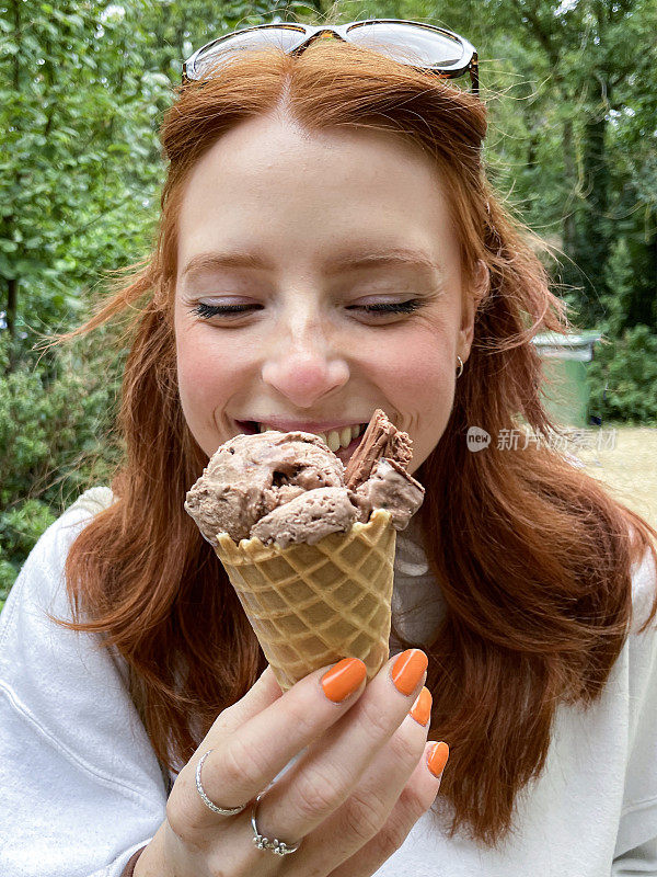 特写照片中，迷人的红发女子头戴墨镜，涂着橙色指甲油，拿着含有巧克力冰淇淋的华夫蛋筒，吃着融化的冰淇淋，背景是林地，前景是焦点