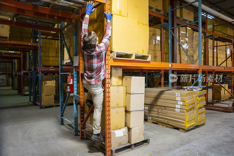 一名年轻的男性仓库工人爬上架子取一个箱子。