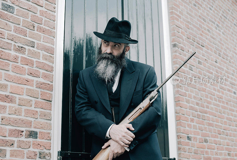 一个戴着帽子留着长胡子的人拿着步枪站在百叶窗前。