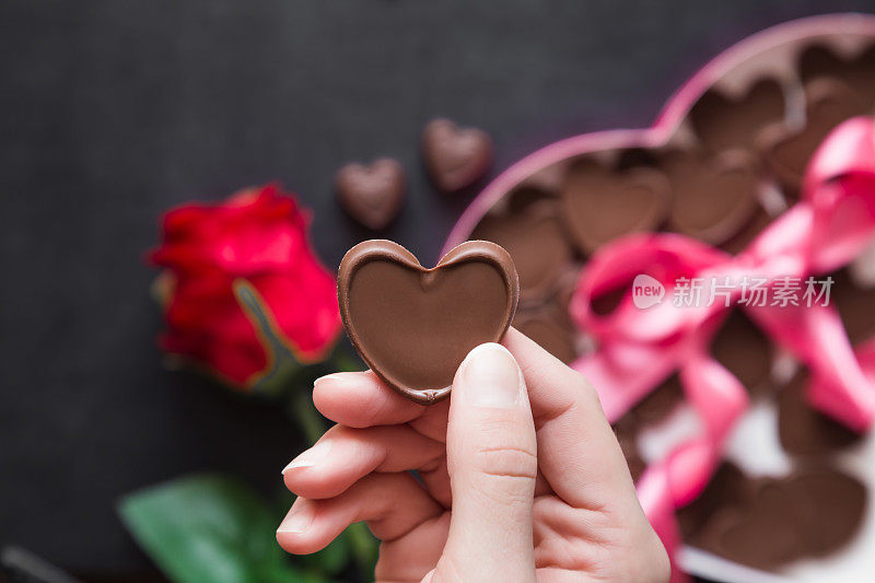女人的手拿着一颗巧克力心。心爱的人送的可爱礼物。桌上放着红玫瑰和带粉色丝带的心形巧克力盒。享受糖果。情人节和关系概念。
