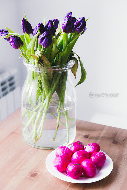 白色的盘子上放着粉色的复活节彩蛋，玻璃花瓶里插着紫色的郁金香，这就是春季复活节的概念