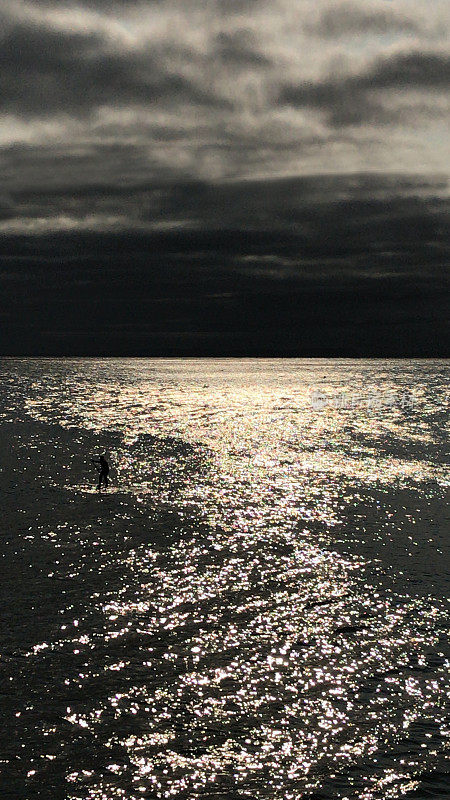 图片中的冲浪者穿着氯丁橡胶潜水衣站在桨板在平静的海面下，乌云密布的天空