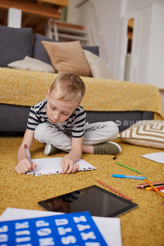 一个注意力集中的金发男生坐在柔软的地毯上，一边写着纸，一边在客厅里做作业