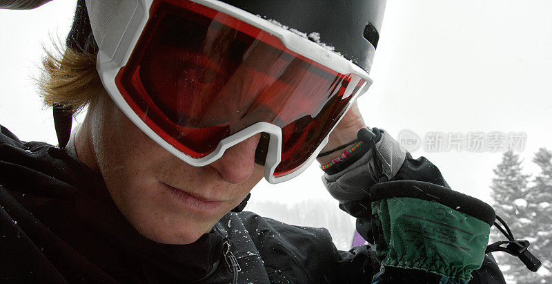 一个20多岁的男性滑雪板运动员戴上他的护目镜，准备在雪上滑雪，阴天。冬季的一天